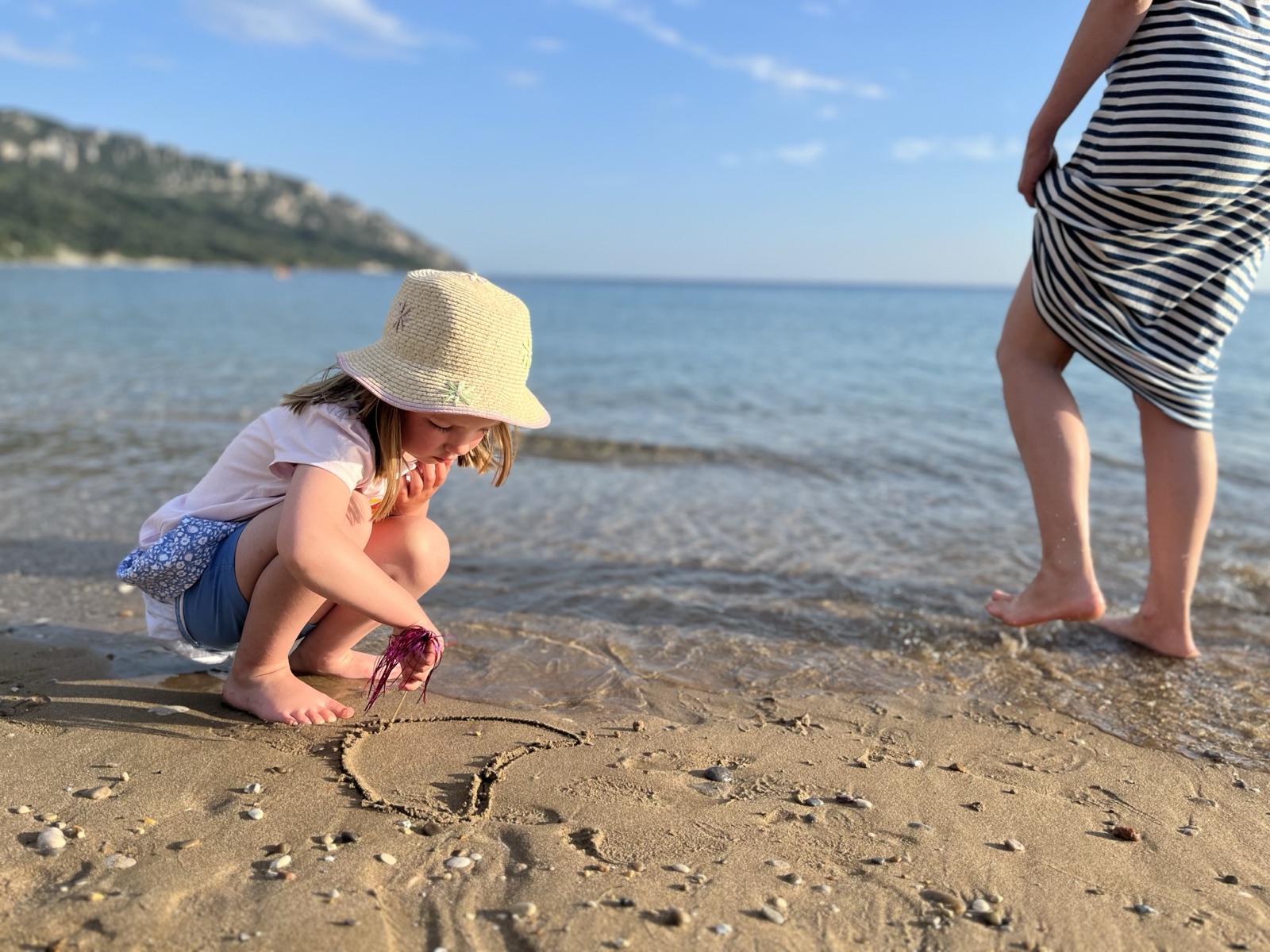 A child in a sun hat drawing a heart in the sand at the edge of the sea on a family holiday
